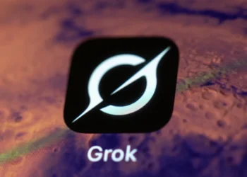 نمای نزدیک از لوگوی سفید Grok روی یک مربع مشکی، در برابر پس‌زمینه‌ای تار و بنفش‌رنگ که بخشی از رابط کاربری یا نمایشگر را نشان می‌دهد.