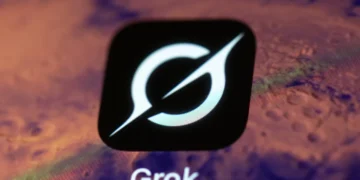 نمای نزدیک از لوگوی سفید Grok روی یک مربع مشکی، در برابر پس‌زمینه‌ای تار و بنفش‌رنگ که بخشی از رابط کاربری یا نمایشگر را نشان می‌دهد.