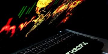 نمای مایل و کوتاهعمق از لپتاپی با صفحه نمایش رنگارنگ و درخشان حاوی نمودارهای بازار مالی، لوگوی شرکت هوش مصنوعی Anthropic روی درپوش آن و محیطی تاریک با نورپردازی نئونی.