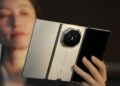 زنی گوشی تاشوی باز Huawei Mate XT 2 در رنگ سفید-طلایی را با دست چپ خود نگه داشته و پشت گوشی با ماژول دوربین هشتضلعی و لوگوی Ultimate Design رو به دوربین است.