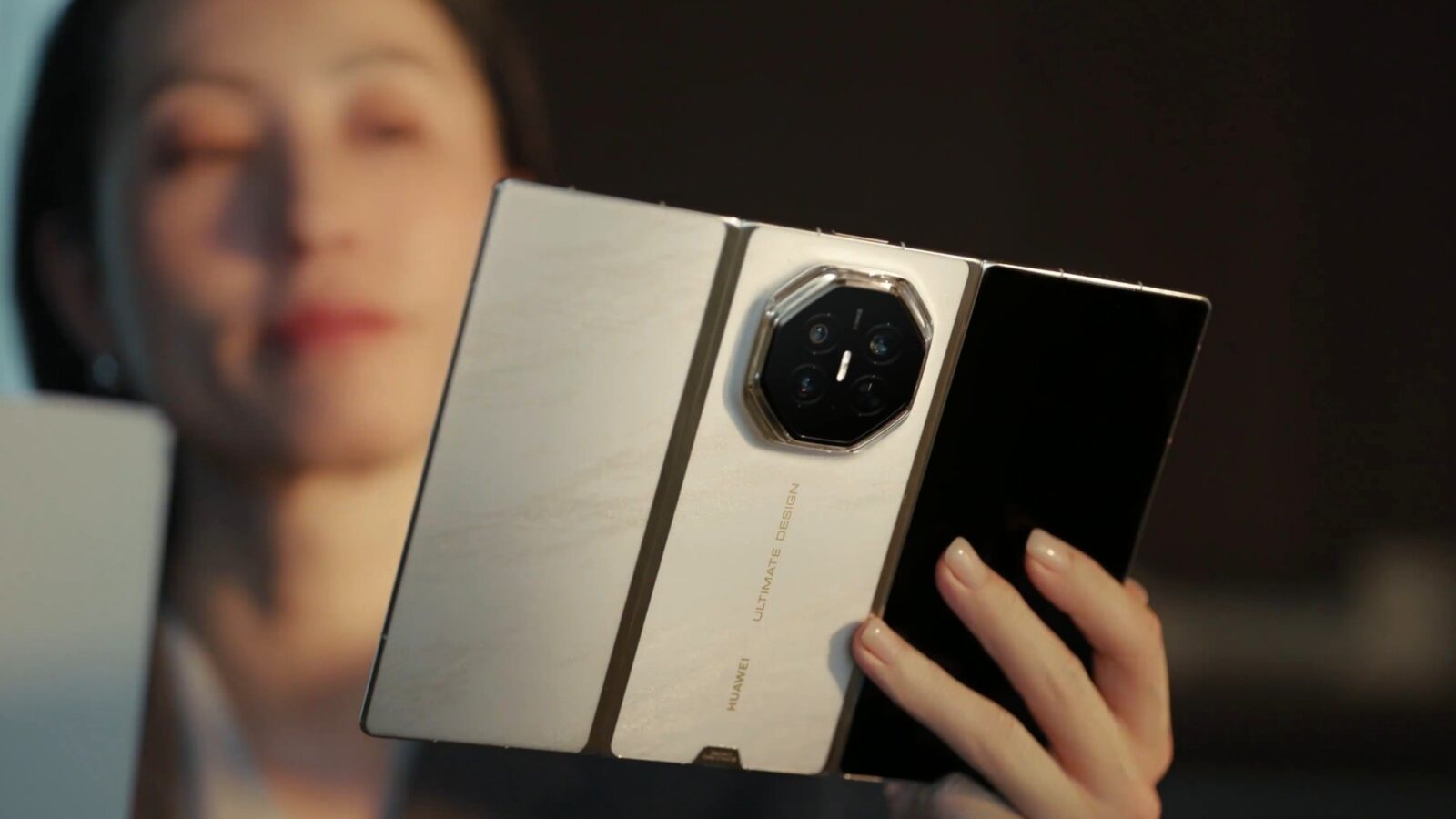 زنی گوشی تاشوی باز Huawei Mate XT 2 در رنگ سفید-طلایی را با دست چپ خود نگه داشته و پشت گوشی با ماژول دوربین هشتضلعی و لوگوی Ultimate Design رو به دوربین است.