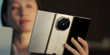 زنی گوشی تاشوی باز Huawei Mate XT 2 در رنگ سفید-طلایی را با دست چپ خود نگه داشته و پشت گوشی با ماژول دوربین هشتضلعی و لوگوی Ultimate Design رو به دوربین است.