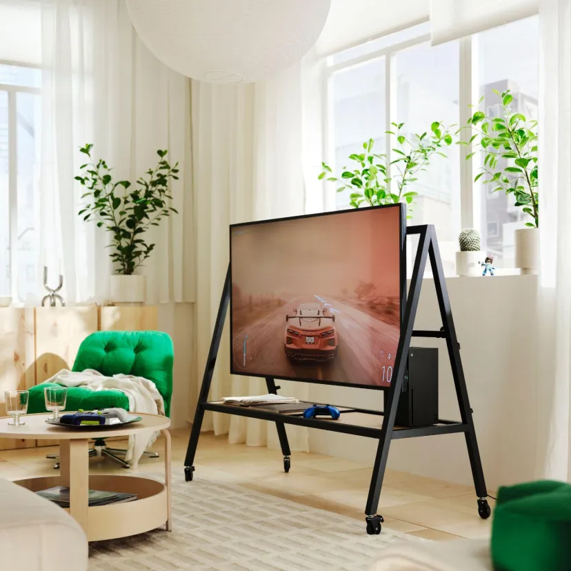 نمایشگر روی پایه مشکی چرخدار مجموعه IKEA و Xbox، در اتاق نشیمنی روشن با تصویر یک خودروی مسابقهای روی صفحه، کنسول بازی و دسته آبی روی طبقه پایینی.