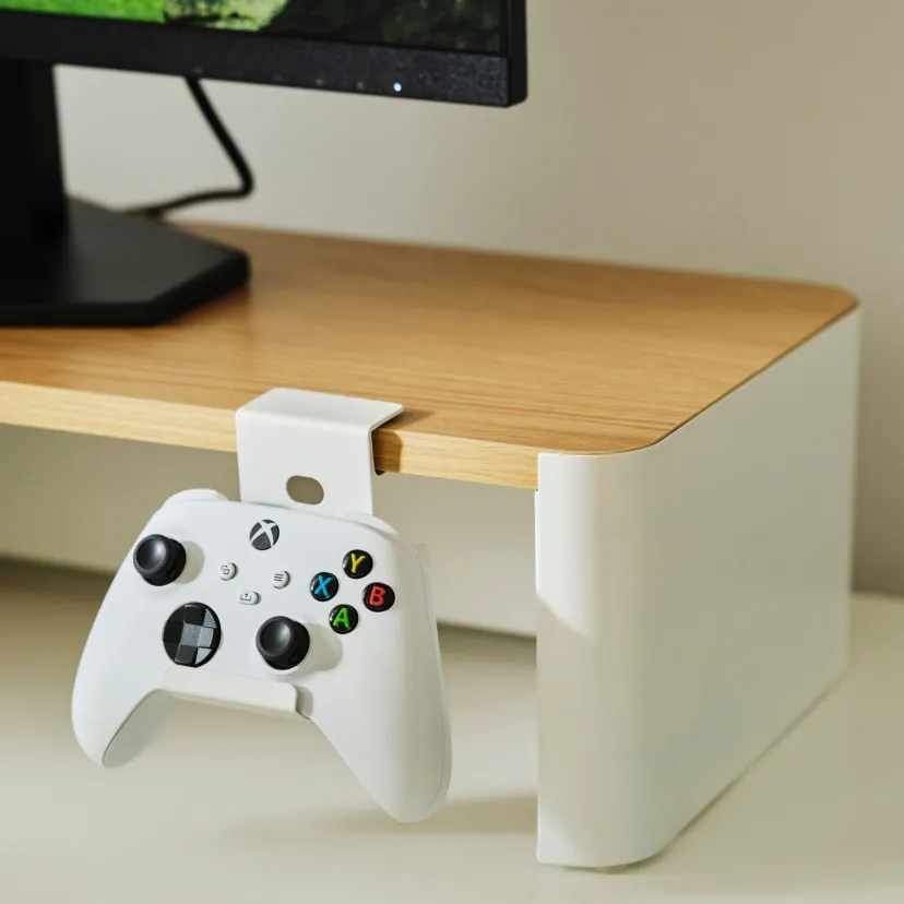 دسته بازی سفید Xbox از قلاب نصبشده در کنار میز چوبی آویزان است. بخشی از نمایشگر و پایه آن در پسزمینه دیده میشود.