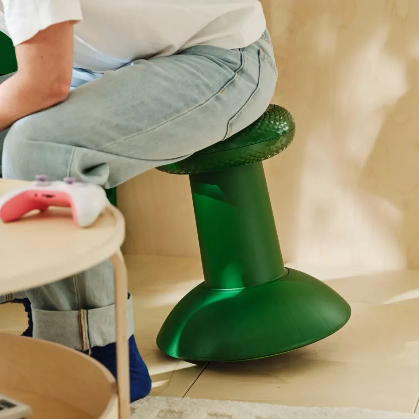 شخصی با شلوار جین روی چهارپایه سبز مجموعه IKEA و Xbox نشسته است. دسته بازی سفید و صورتی روی میز کوچک کنار چهارپایه دیده میشود.