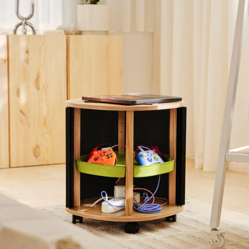 میز کناری مشکی و چوبی مجموعه IKEA و Xbox با چرخهای کوچک، محفظههای داخلی دارای بندهای سبز برای نگهداری دستههای بازی رنگارنگ و شارژرها، و یک لپتاپ روی سطح بالایی.