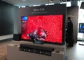 یک تلویزیون بزرگ ۶۵ اینچی سامسونگ با فناوری Micro RGB در یک نمایشگاه یا فروشگاه که تصویری بسیار درخشان و زنده با غلبه رنگ قرمز شدید شامل یک فانوس دریایی قرمز در میان هزاران گوی قرمز کوچک را نشان میدهد. مردی با کتوشلوار تیره در سمت راست تلویزیون ایستاده و لوگوی سامسونگ در پسزمینه چپ دیده میشود.