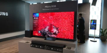 یک تلویزیون بزرگ ۶۵ اینچی سامسونگ با فناوری Micro RGB در یک نمایشگاه یا فروشگاه که تصویری بسیار درخشان و زنده با غلبه رنگ قرمز شدید شامل یک فانوس دریایی قرمز در میان هزاران گوی قرمز کوچک را نشان میدهد. مردی با کتوشلوار تیره در سمت راست تلویزیون ایستاده و لوگوی سامسونگ در پسزمینه چپ دیده میشود.
