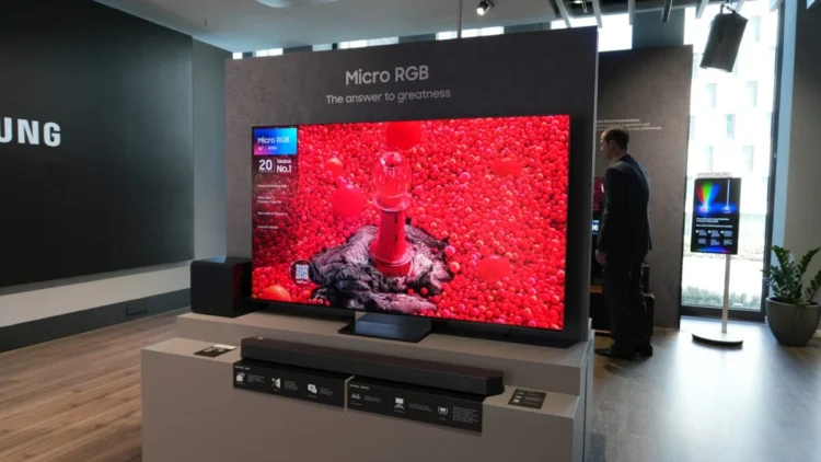 یک تلویزیون بزرگ ۶۵ اینچی سامسونگ با فناوری Micro RGB در یک نمایشگاه یا فروشگاه که تصویری بسیار درخشان و زنده با غلبه رنگ قرمز شدید شامل یک فانوس دریایی قرمز در میان هزاران گوی قرمز کوچک را نشان میدهد. مردی با کتوشلوار تیره در سمت راست تلویزیون ایستاده و لوگوی سامسونگ در پسزمینه چپ دیده میشود.