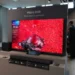 یک تلویزیون بزرگ ۶۵ اینچی سامسونگ با فناوری Micro RGB در یک نمایشگاه یا فروشگاه که تصویری بسیار درخشان و زنده با غلبه رنگ قرمز شدید شامل یک فانوس دریایی قرمز در میان هزاران گوی قرمز کوچک را نشان میدهد. مردی با کتوشلوار تیره در سمت راست تلویزیون ایستاده و لوگوی سامسونگ در پسزمینه چپ دیده میشود.