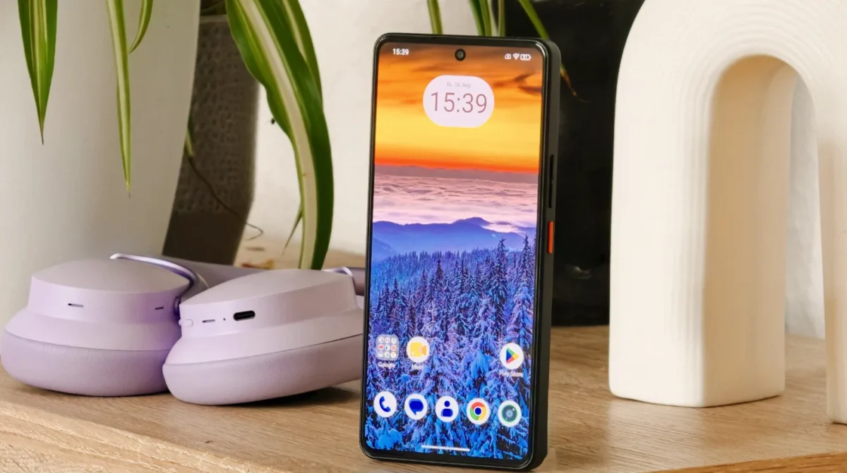 نمای روبهروی گوشی گیگاست GS6 Pro با والپیپر طبیعت برفی در کنار یک هدفون بنفش روی میز چوبی.