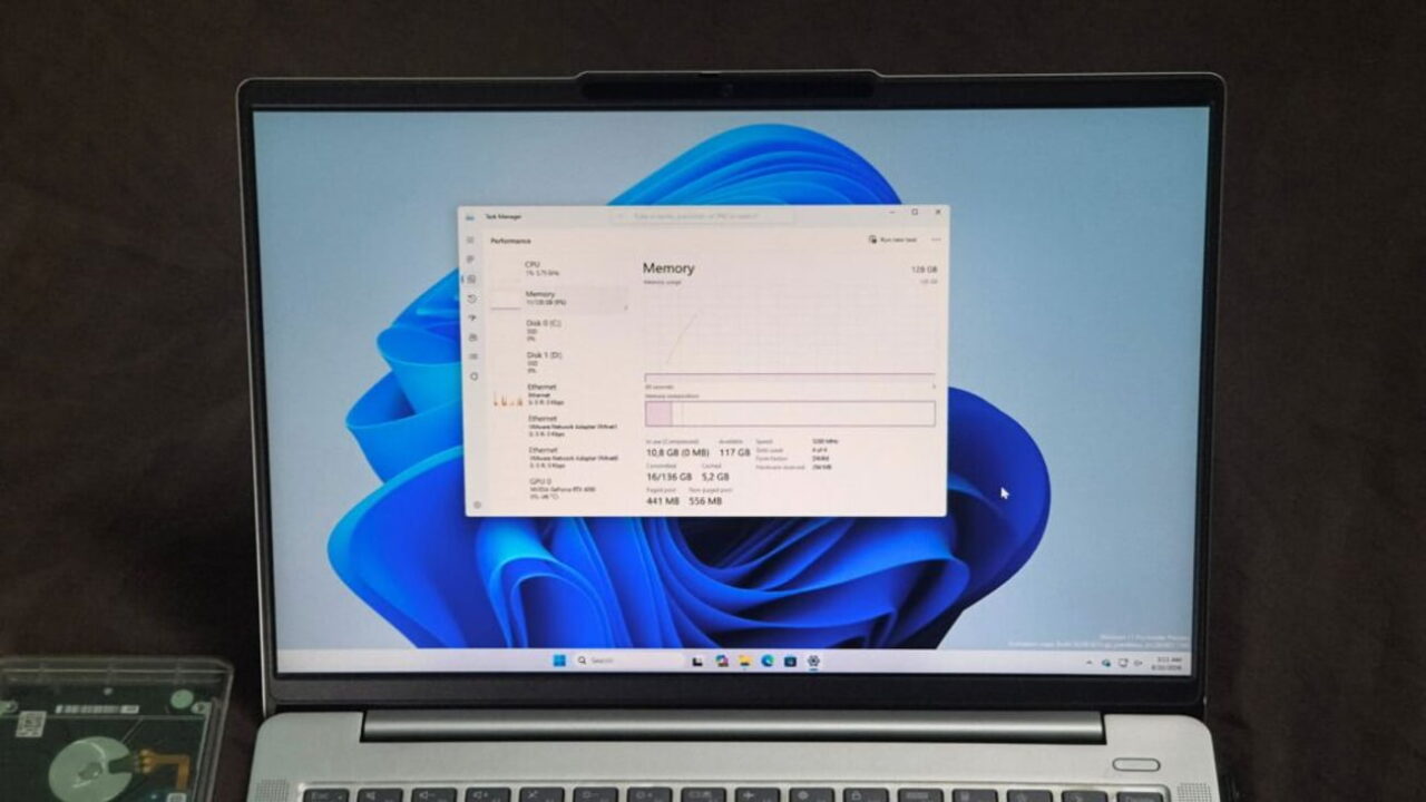 نمای روبرو از نمایشگر لپتاپ نقرهای با والپیپر آبی ویندوز ۱۱ و پنجره باز Task Manager در تب Performance، که بخش Memory با ظرفیت کل ۱۲۸ گیگابایت را نشان میدهد.