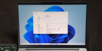 نمای روبرو از نمایشگر لپتاپ نقرهای با والپیپر آبی ویندوز ۱۱ و پنجره باز Task Manager در تب Performance، که بخش Memory با ظرفیت کل ۱۲۸ گیگابایت را نشان میدهد.