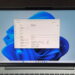 نمای روبرو از نمایشگر لپتاپ نقرهای با والپیپر آبی ویندوز ۱۱ و پنجره باز Task Manager در تب Performance، که بخش Memory با ظرفیت کل ۱۲۸ گیگابایت را نشان میدهد.