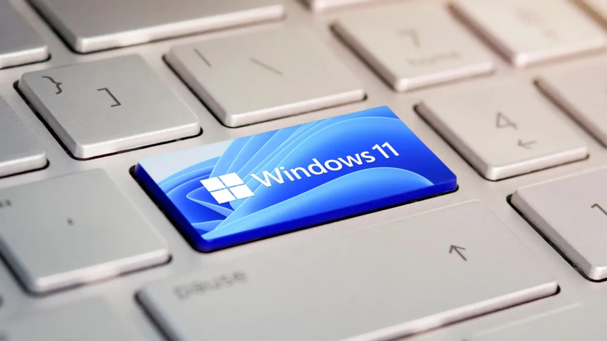 کلید آبی رنگ Windows 11 با لوگوی ویندوز و پس زمینه موجی، میان کلیدهای نقره ای یک کیبورد.
