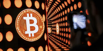 نمایشگر بزرگ نورانی با لوگوی نارنجی بیتکوین در یک راهروی تونلی با چراغهای دایرهای نارنجی و بازتابهای بیپایان.