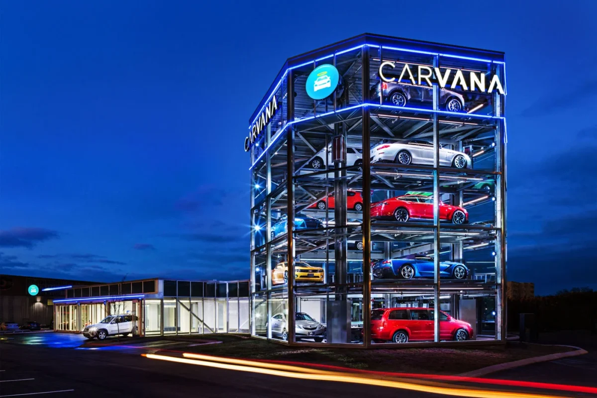 نمای زاویهدار از دستگاه فروش خودروی شیشهای Carvana با چندین خودروی پارکشده در طبقات و نورپردازی آبی در شب