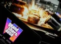 صفحه نمایش گوشی هوشمند با نمایش لوگوی نئونی Grand Theft Auto VI و تاریخ انتشار ۲۶ می ۲۰۲۶ در مقابل مانیتور پخش گیمپلی تصادف خودروها.
