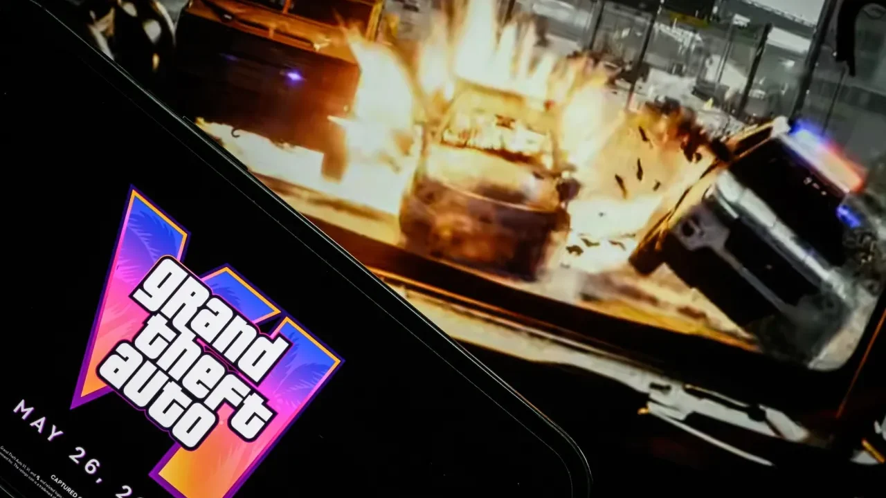 صفحه نمایش گوشی هوشمند با نمایش لوگوی نئونی Grand Theft Auto VI و تاریخ انتشار ۲۶ می ۲۰۲۶ در مقابل مانیتور پخش گیمپلی تصادف خودروها.