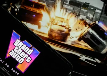صفحه نمایش گوشی هوشمند با نمایش لوگوی نئونی Grand Theft Auto VI و تاریخ انتشار ۲۶ می ۲۰۲۶ در مقابل مانیتور پخش گیم‌پلی تصادف خودروها.