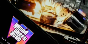 صفحه نمایش گوشی هوشمند با نمایش لوگوی نئونی Grand Theft Auto VI و تاریخ انتشار ۲۶ می ۲۰۲۶ در مقابل مانیتور پخش گیمپلی تصادف خودروها.