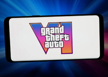 لوگوی Grand Theft Auto VI روی نمایشگر سفید یک گوشی هوشمند، با پس‌زمینه‌ای از پرتوهای نور آبی، صورتی و قرمز.