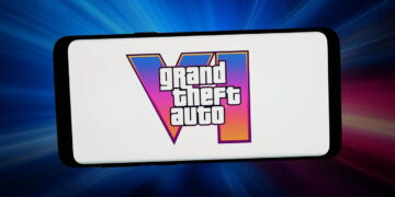 لوگوی Grand Theft Auto VI روی نمایشگر سفید یک گوشی هوشمند، با پسزمینهای از پرتوهای نور آبی، صورتی و قرمز.