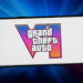 لوگوی Grand Theft Auto VI روی نمایشگر سفید یک گوشی هوشمند، با پسزمینهای از پرتوهای نور آبی، صورتی و قرمز.