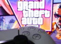 کنترلر مشکی کنسول در پیشزمینه و لوگوی بزرگ Grand Theft Auto با تصاویر رنگارنگ شخصیتها و فضای ساحلی GTA 6 در پسزمینه.