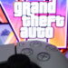 کنترلر مشکی کنسول در پیشزمینه و لوگوی بزرگ Grand Theft Auto با تصاویر رنگارنگ شخصیتها و فضای ساحلی GTA 6 در پسزمینه.