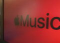 نمای نزدیک از لوگوی Apple Music با نماد اپل و عبارت Music روی پس‌زمینه‌ای قرمز و نورپردازی‌شده.