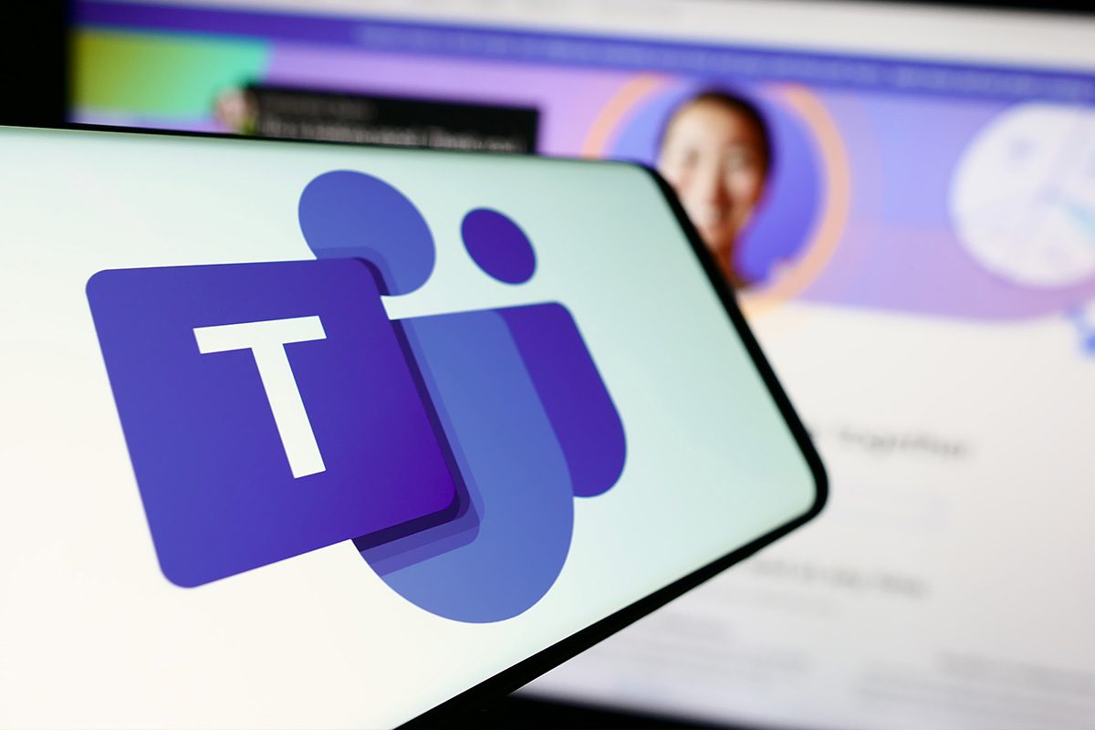 نمای نزدیک از گوشی هوشمند با لوگوی Microsoft Teams روی صفحه، در حالی که پس‌زمینه‌ای محو از رابط کاربری تماس یا جلسه آنلاین روی نمایشگر دیده می‌شود.