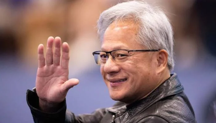 جنسن هوانگ (Jensen Huang)، مدیرعامل شرکت انویدیا، با ظاهر شناختهشدهاش شامل موهای نقرهای و عینک، در حالی که کت چرمی مشکی به تن دارد و در یک رویداد رسمی برای مخاطبان دست تکان میدهد.