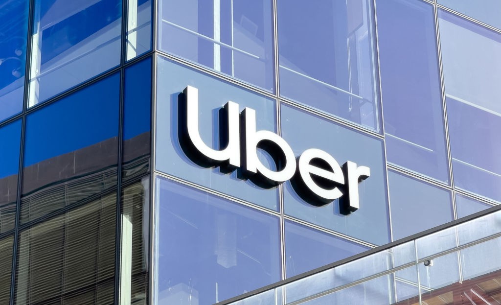نمای نزدیک از تابلوی فلزی برجسته با لوگوی سفید اوبر (Uber) روی نمای شیشه‌ای مدرن ساختمان مرکزی این شرکت در نور روز.
