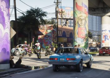 صحنه‌ای از بازی GTA 6 با ماشین کلاسیک آبی، موتورسواران در حال تک‌چرخ زدن و ستون‌های نقاشی‌شده پل در وایس سیتی.