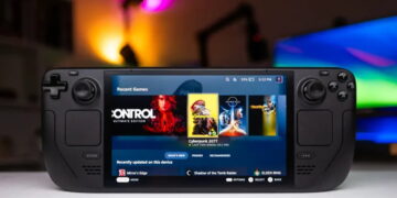 کنسول بازی دستی Steam Deck مشکی با نمایشگر روشن در حال اجرای منوی بازی‌ها شامل Cyberpunk 2077 روی میز