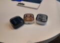 سه کیس شارژ هدفون بیسیم سامسونگ Galaxy Buds در رنگهای مشکی، کرم و نقرهای با درپوش نیمهشفاف دودی روی میز سفید در کنار یک گوشی هوشمند