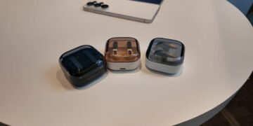 سه کیس شارژ هدفون بیسیم سامسونگ Galaxy Buds در رنگهای مشکی، کرم و نقرهای با درپوش نیمهشفاف دودی روی میز سفید در کنار یک گوشی هوشمند