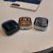 سه کیس شارژ هدفون بیسیم سامسونگ Galaxy Buds در رنگهای مشکی، کرم و نقرهای با درپوش نیمهشفاف دودی روی میز سفید در کنار یک گوشی هوشمند