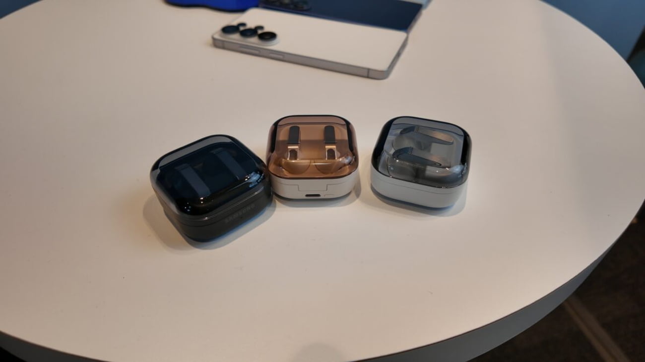 سه کیس شارژ هدفون بیسیم سامسونگ Galaxy Buds در رنگهای مشکی، کرم و نقرهای با درپوش نیمهشفاف دودی روی میز سفید در کنار یک گوشی هوشمند