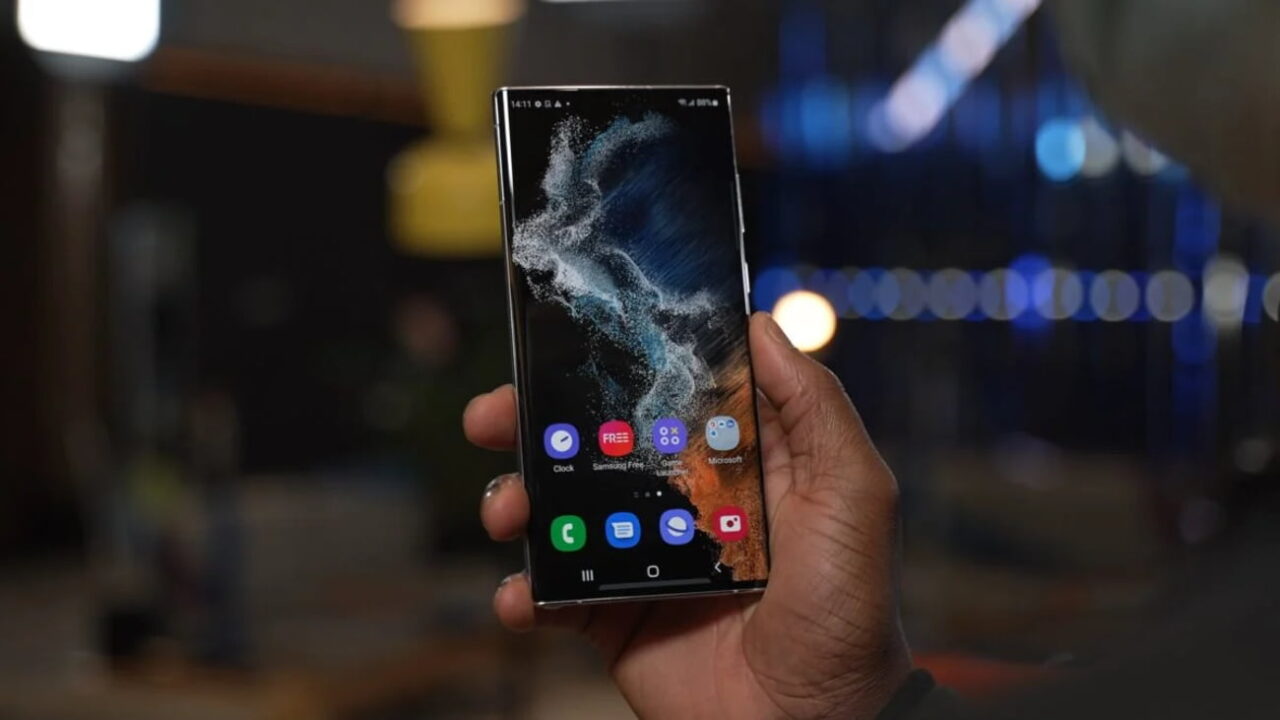 گوشی سامسونگ گلکسی اس ۲۲ اولترا با نمایشگر روشن و تصویر پسزمینه انتزاعی، در دست کاربر و مقابل پسزمینهای تاریک با نورهای محو.