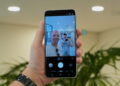 نمای دستی که گوشی Samsung Galaxy S26 FE را در حالت Photo گرفته؛ صفحه نمایش پیش‌نمایش دوربین، دو نفر در محیط صنعتی روشن و چند دکمه‌ی تنظیمات از قبیل Portrait و Video را نشان می‌دهد.