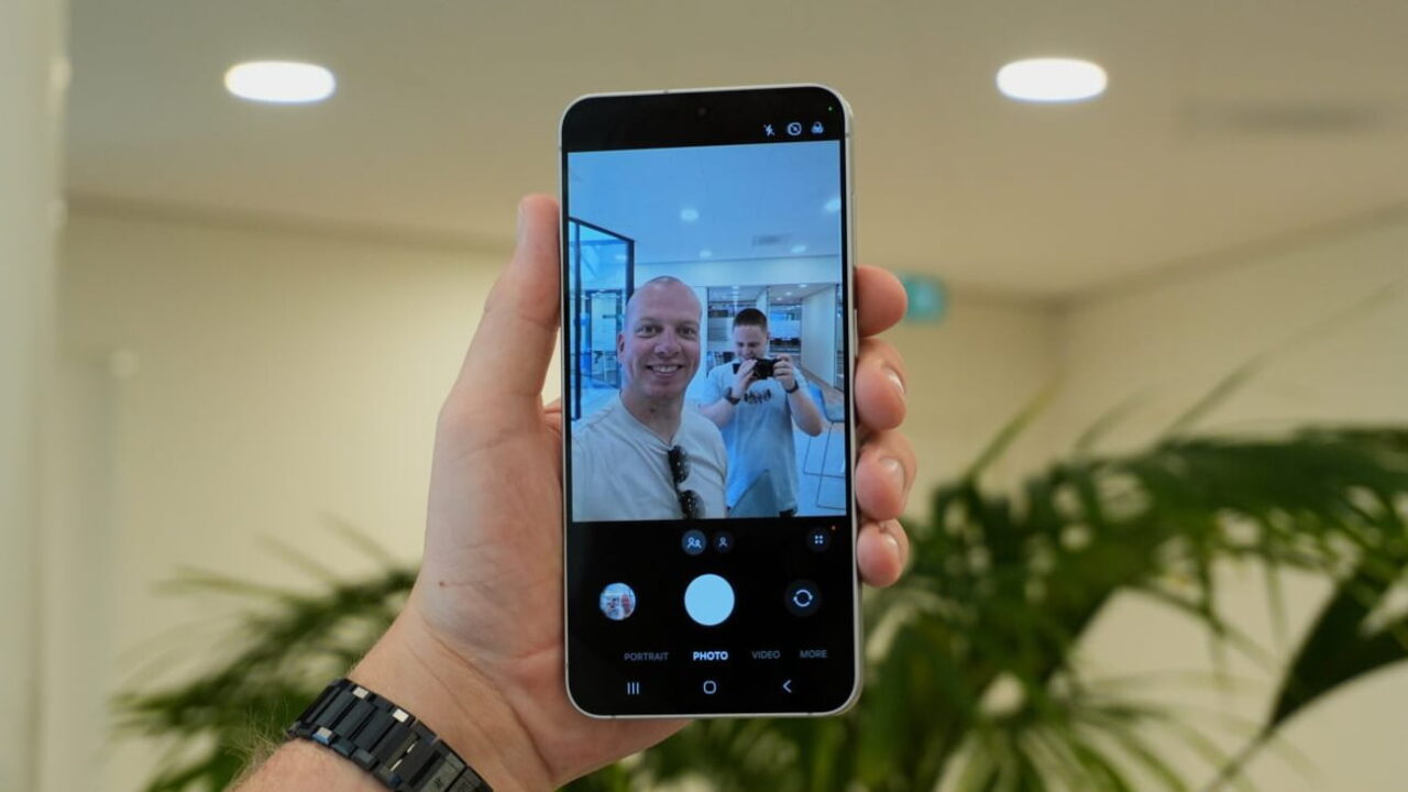 نمای دستی که گوشی Samsung Galaxy S26 FE را در حالت Photo گرفته؛ صفحه نمایش پیشنمایش دوربین، دو نفر در محیط صنعتی روشن و چند دکمهی تنظیمات از قبیل Portrait و Video را نشان میدهد.