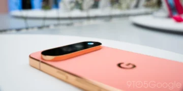 عکس نزدیک از پشت گوشی صورتی Pixel 11 که روی میز سفید قرار گرفته. ماژول دوربین افقی با قاب طلایی روی بدنه دیده می‌شود و چراغ کوچکی به رنگ آبی در انتهای آن می‌درخشد. لوگوی G گوگل روی بدنه و واترمارک 9to5Google در گوشه پایین تصویر است.