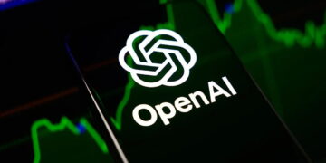 گوشی هوشمند با لوگوی سفید و نام OpenAI روی صفحه نمایش در مقابل پس‌زمینه سبز و تیره نمودارهای بازارهای مالی.