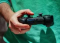 نمای نزدیک از دست فردی که یک کنترلر مشکی سونی DualShock 3 با برند SIXAXIS را در دست دارد، در پسزمینهای با لباس سبز
