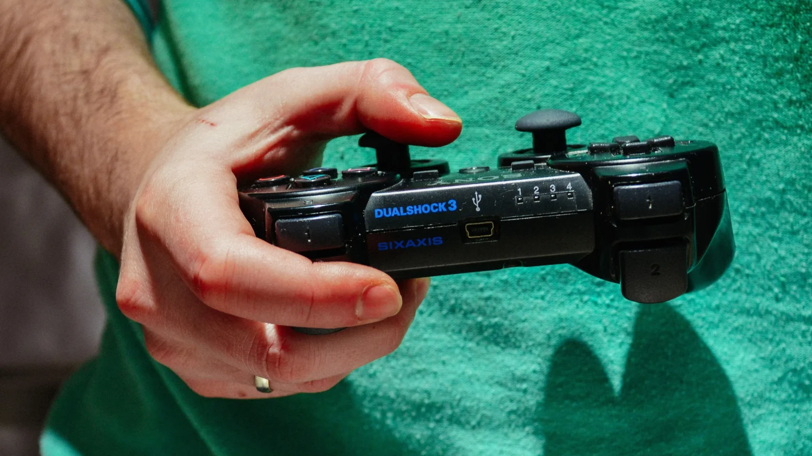 نمای نزدیک از دست فردی که یک کنترلر مشکی سونی DualShock 3 با برند SIXAXIS را در دست دارد، در پسزمینهای با لباس سبز