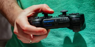 نمای نزدیک از دست فردی که یک کنترلر مشکی سونی DualShock 3 با برند SIXAXIS را در دست دارد، در پسزمینهای با لباس سبز