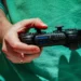 نمای نزدیک از دست فردی که یک کنترلر مشکی سونی DualShock 3 با برند SIXAXIS را در دست دارد، در پسزمینهای با لباس سبز