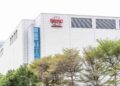 نمای بیرونی ساختمان مدرن کارخانه تولید نیمههادی شرکت TSMC با نمای سفید و پنجرههای شیشهای آبیرنگ، با لوگوی قرمز TSMC در بالای سازه و ردیفی از درختان سرسبز در پیشزمینه.